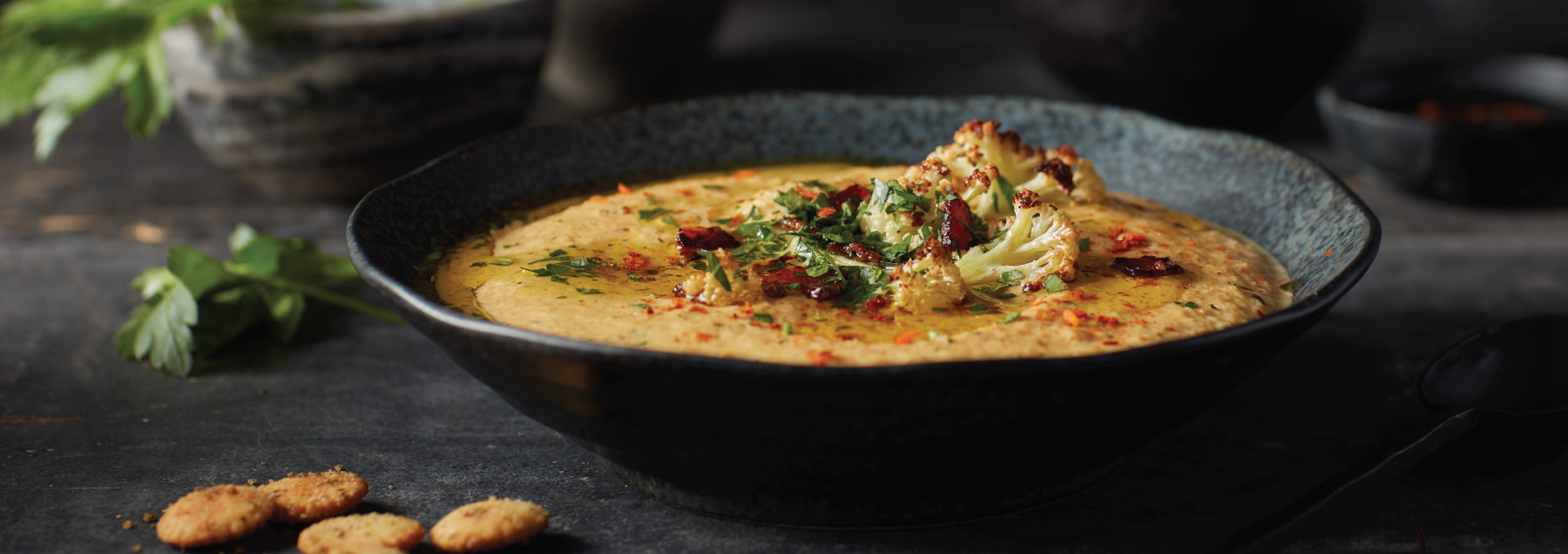 creamy cauliflower soup topped with cauliflower and herbs in a black bowl on a black table with crackers and herbs on the side