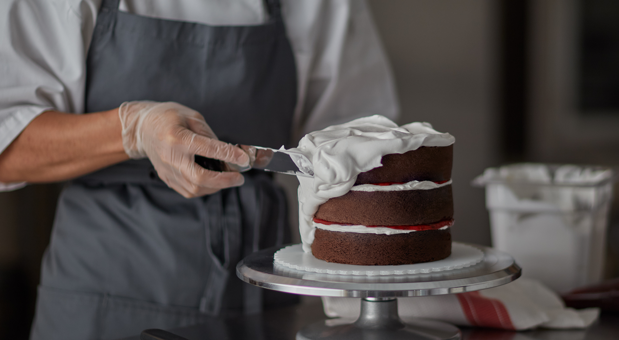 baker icing a chocolate layer cake in a bakery back of house